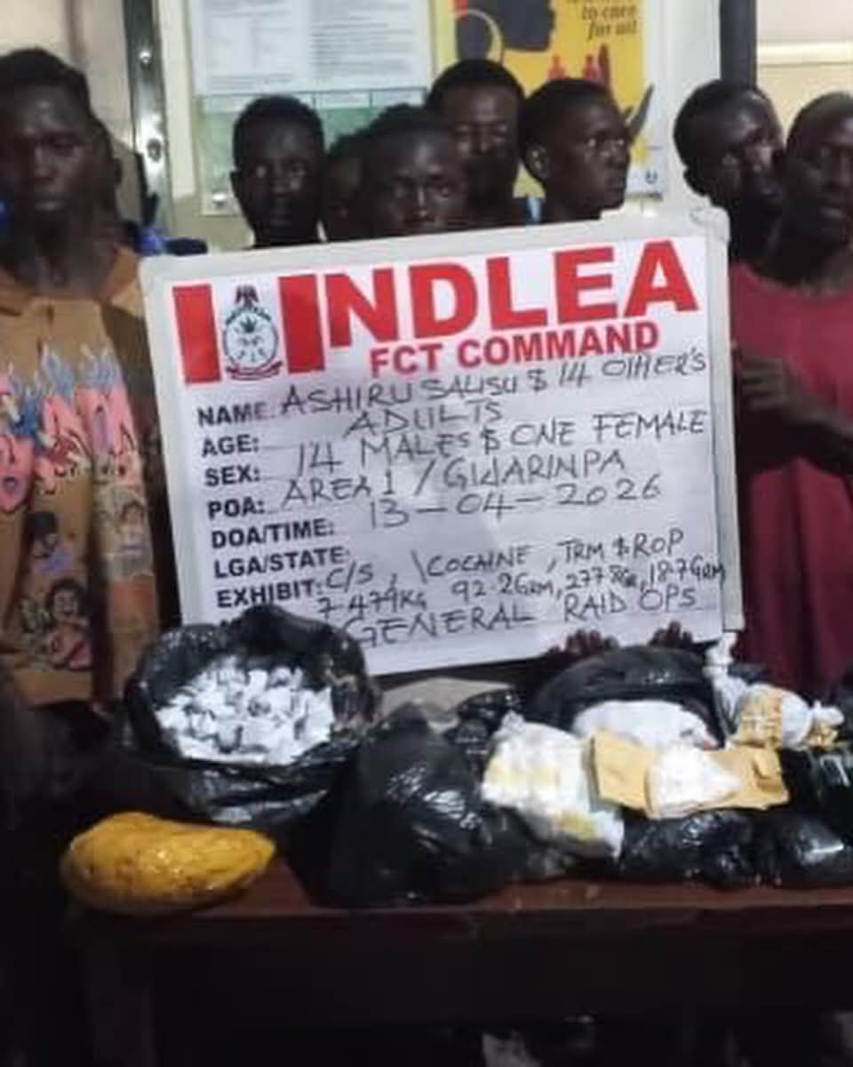 People standing behind a large poster with bold red text, containing handwritten case details; bags and packages sit on a table in front.