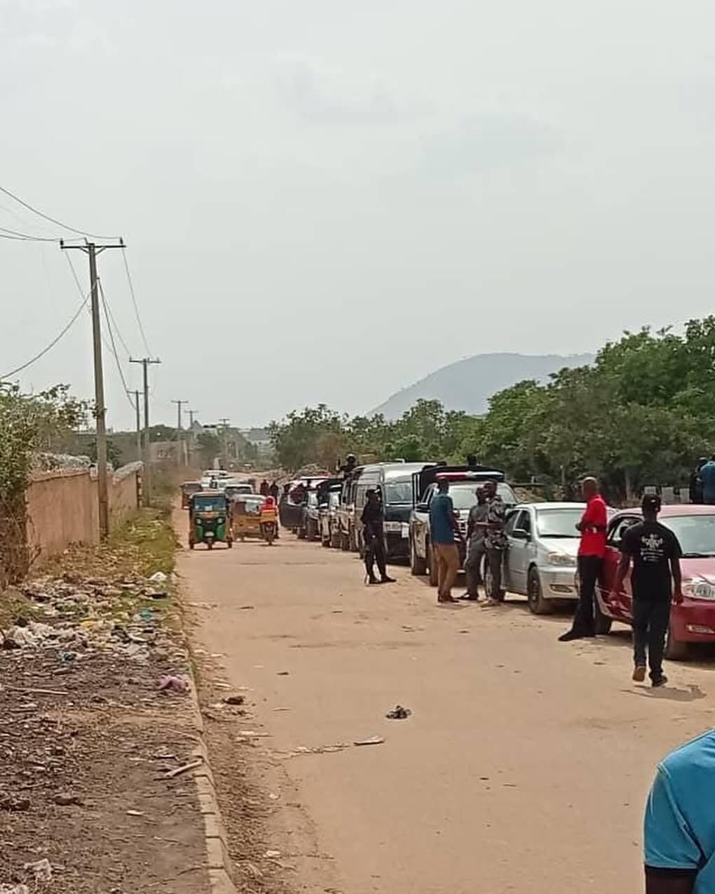 Line of parked cars along a dusty rural street with people standing nearby and utility poles running along the road.