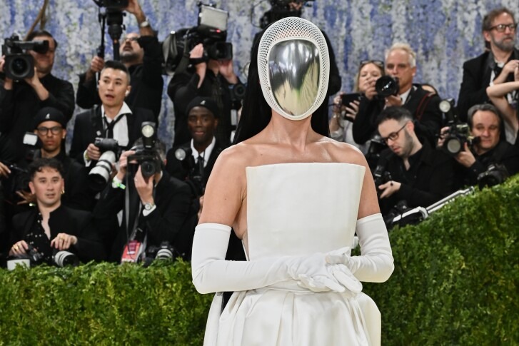 Anna Wintour (Daily Express) - THEWILL NEWS MEDIA Person in a white strapless gown and long white gloves posing on a red carpet with a reflective metallic mask, surrounded by photographers.