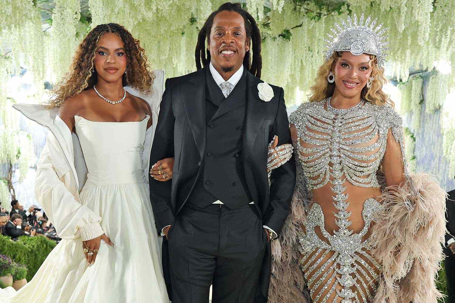 Blue Ivy, Jay Z, Beyoncé (People.com) - THEWILL NEWS MEDIA Three well-dressed individuals posing together at a formal event: a woman in a white off-the-shoulder gown on the left, a man in a black suit and vest center, and a woman in an ornate silver embellished outfit with a crown-like headpiece on the right.