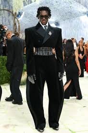 Wisdom Kaye (Hypebeast) - THEWILL NEWS MEDIA Man in an all-black, cape-style formal outfit with a wide metallic belt, gloves, and sunglasses at a red-carpet event.