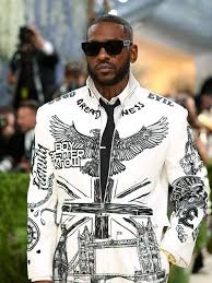 Skepta (Instagram) - THEWILL NEWS MEDIA Man wearing a white graphic jacket with wings and sword designs, sunglasses, at an outdoor fashion event.