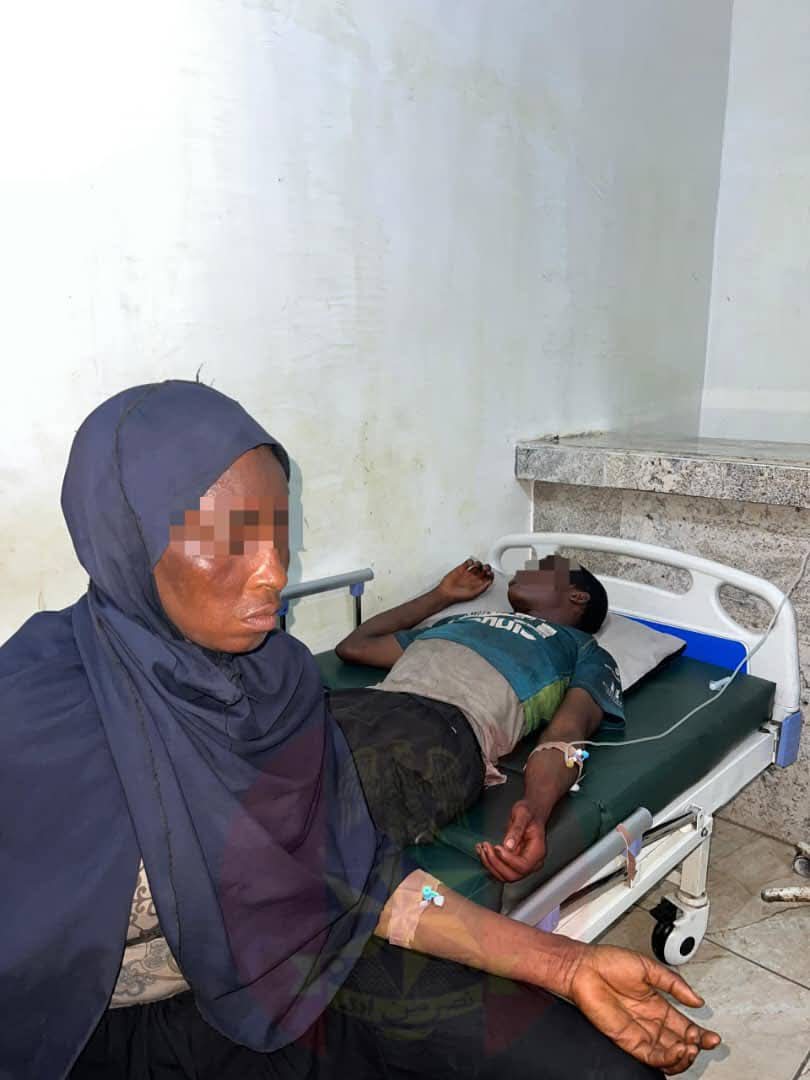 Woman wearing a dark blue hijab sits beside a hospital bed where a man lies, connected to an IV drip in his left arm.