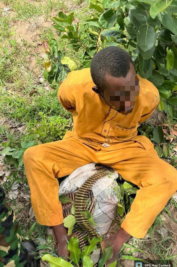 Person in an orange jumpsuit sits outdoors on a wrapped bundle or sack, a long ammunition belt draped over the bundle; the face is pixelated and green plants surround them.