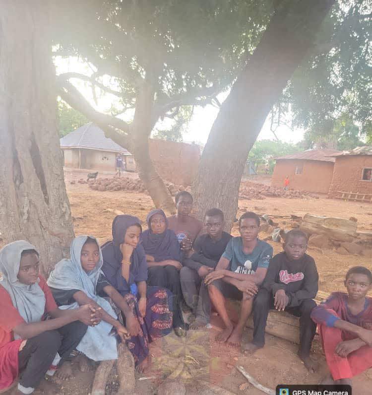 Group of children sitting on a log under a large tree in a dusty village yard, with huts in the background, relaxing and talking together.