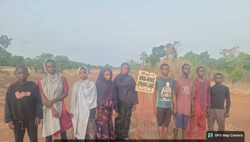 Group of children standing in a dusty rural field, some wearing headscarves, posing for the photo with a handwritten sign held by two kids in the center (GPS watermark bottom right).