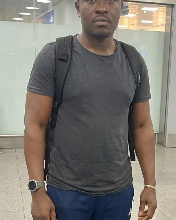 Man wearing a dark gray T-shirt and black backpack standing in an indoor public space, wearing a smartwatch on his left wrist and a gold bracelet on his right wrist.