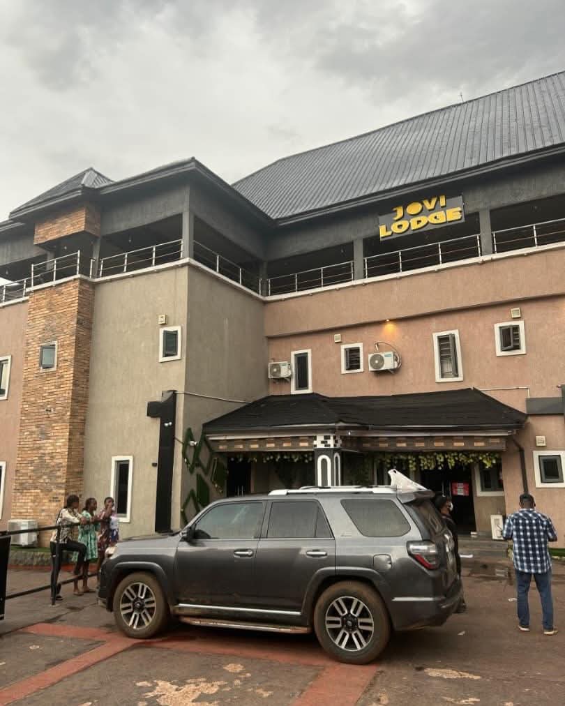 Front view of Jovi Lodge with a gray SUV parked in the driveway and a group of people standing near the entrance on a cloudy day.