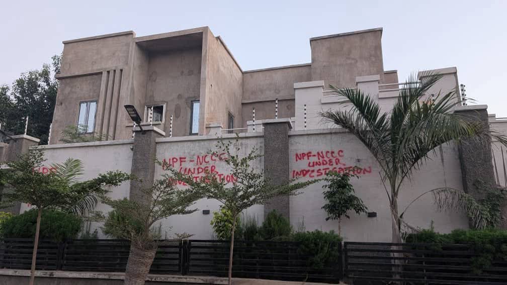 A modern concrete house with a high wall and a palm tree in front; red graffiti on the wall suggesting something about an investigation.