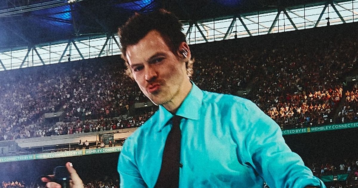 Male performer in a turquoise shirt and black tie taking a selfie on stage with a large crowd behind him in a stadium.