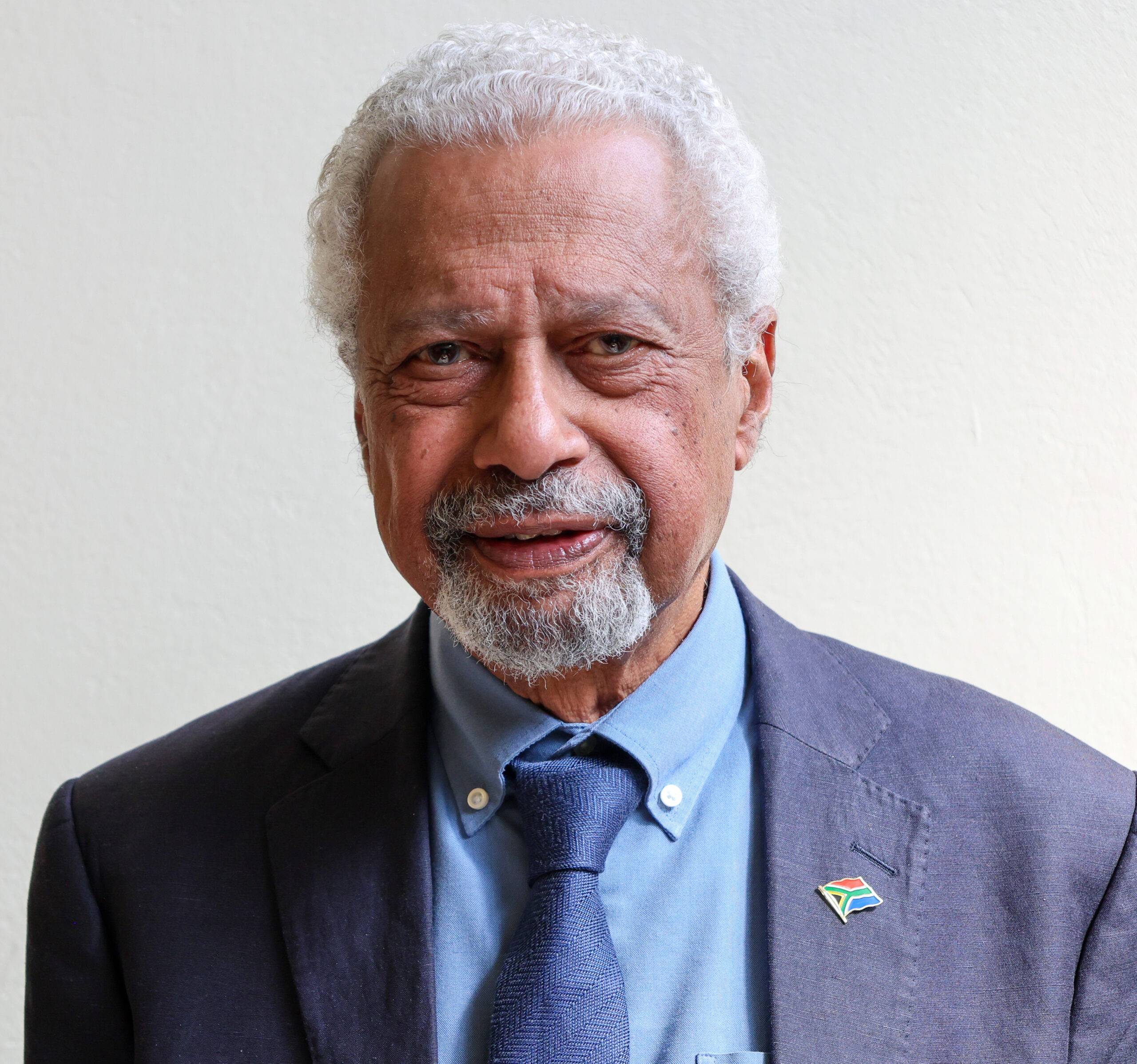 Abdulrazak Gurnah - THEWILL NEWS MEDIA Close-up portrait of an older man with white hair and beard, wearing a dark blazer, blue shirt, and tie, with a South Africa flag lapel pin visible on the lapel.