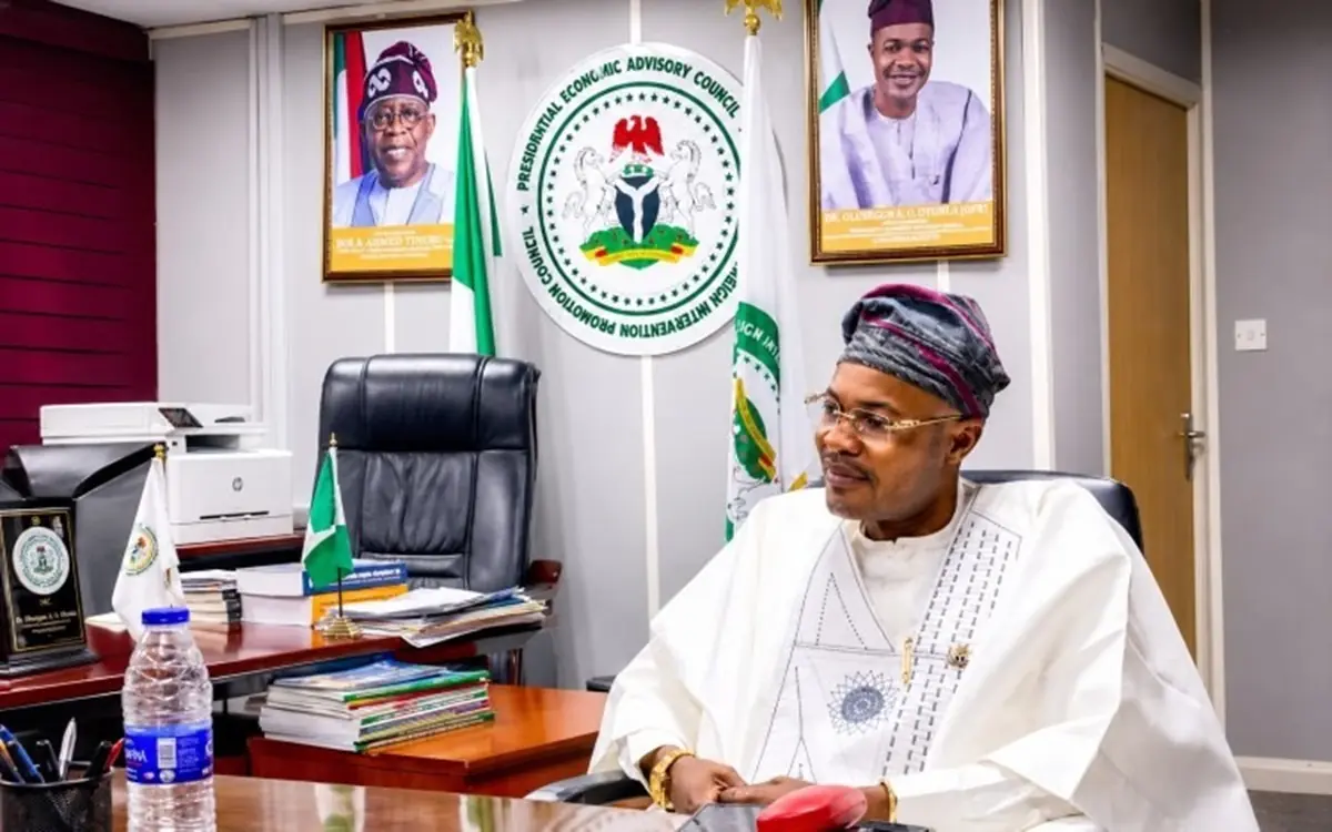 Adeniyi Adeyemi - THEWILL NEWS MEDIA Man in traditional Nigerian attire sits at a desk in a government office with flags and portraits in the background, looking to the side.