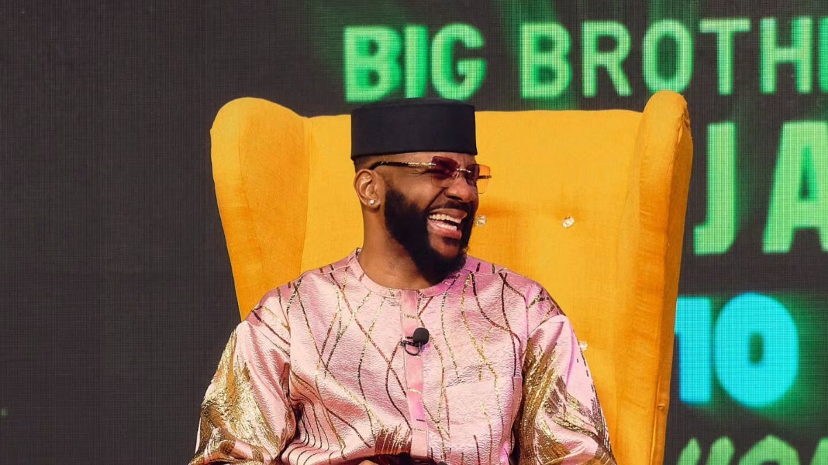 Bearded man in a pink traditional outfit and black cap laughing while seated in a bright yellow high-back chair on a Big Brother Naija stage, mic clipped to his chest.