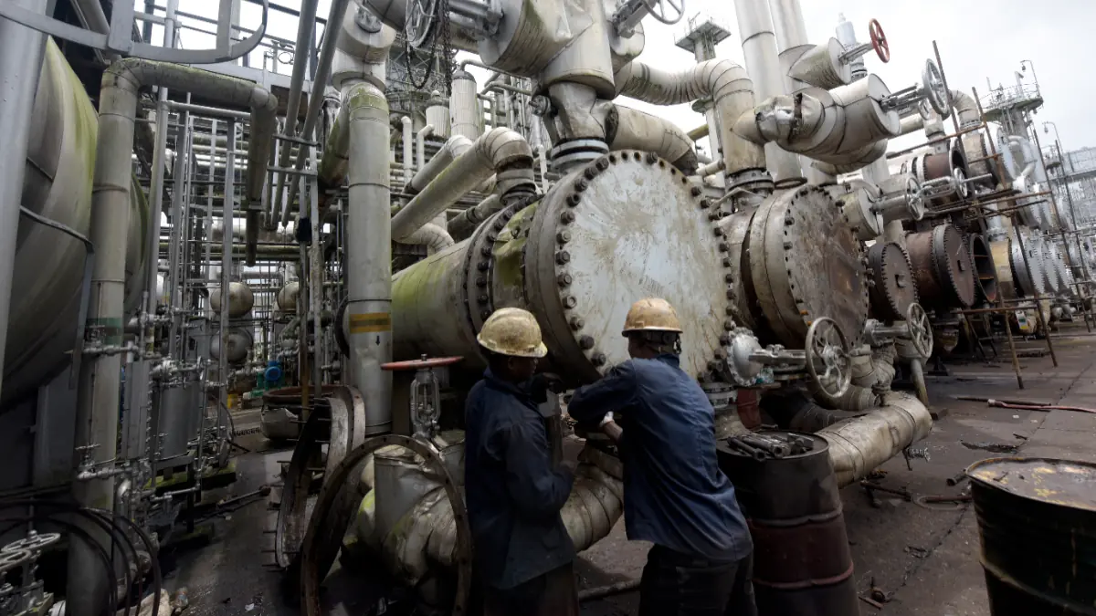 Port Harcourt Refinery - THEWILL NEWS MEDIA Two workers in hard hats inspect a large valve amid a dense network of industrial pipes and equipment at an oil/refinery facility.