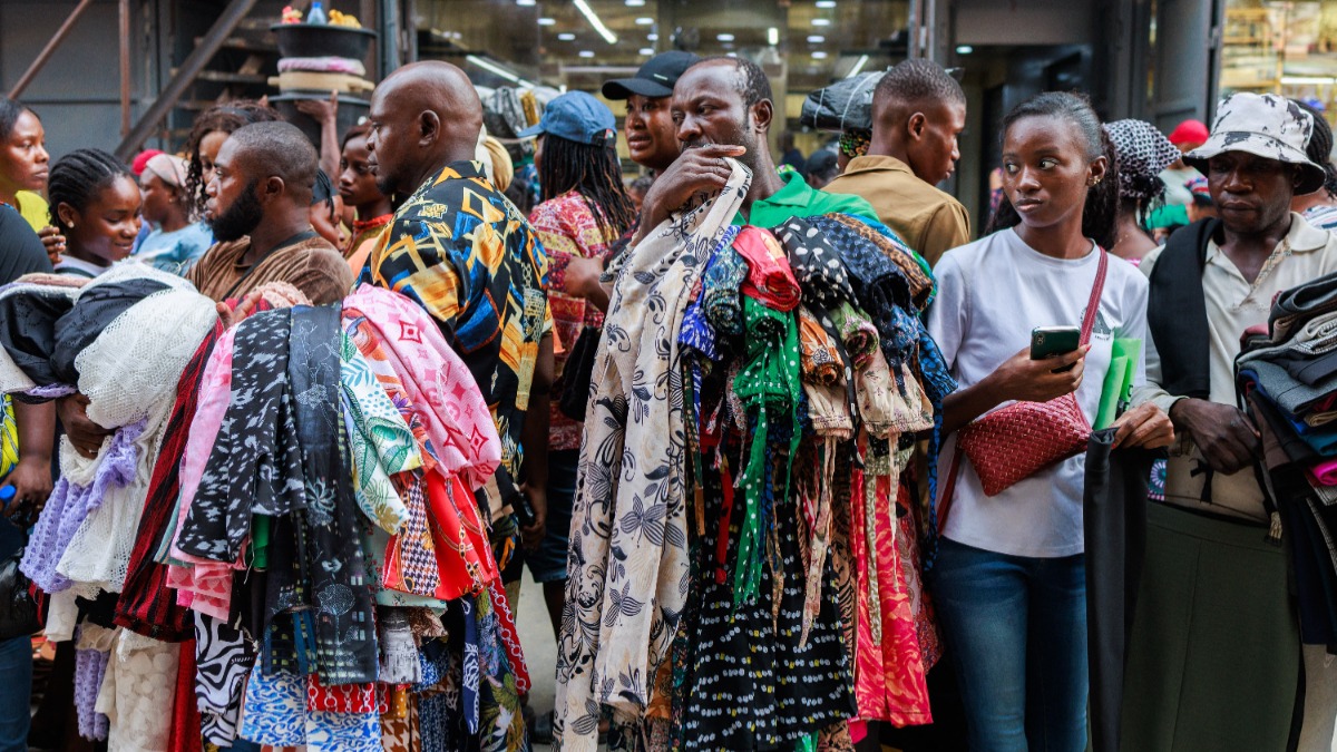 Crowded street market with shoppers holding and examining stacks of colorful fabrics in a textile stall flood of cloths nearby.