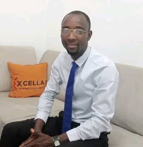 Man in a light blue shirt and blue tie sits on a beige sofa in an office lounge, with an orange cushion in the background.