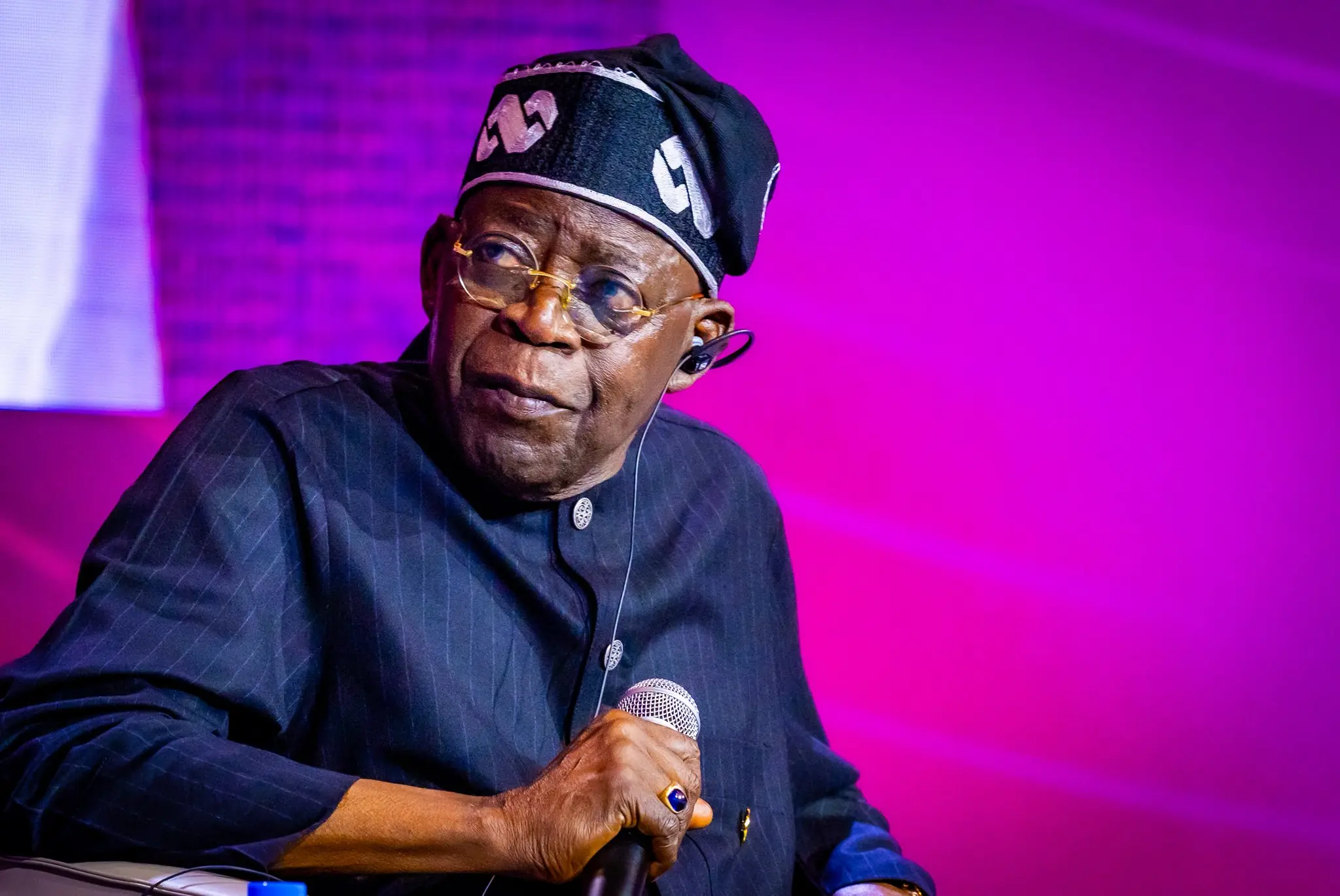 Tinubu - THEWILL NEWS MEDIA Male performer on stage, wearing a dark pinstripe outfit and a head wrap, holding a microphone under purple lighting