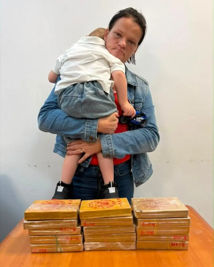 Mother in a denim jacket holds a baby while standing behind a table with stacked brown packages on it, indoors.