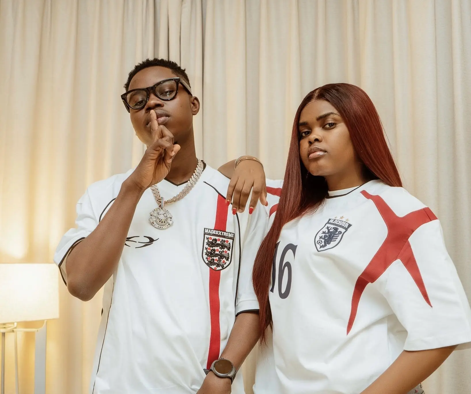 Two people in England football jerseys pose indoors: the man with glasses makes a shushing gesture, the woman with long red hair rests her arm on his shoulder.