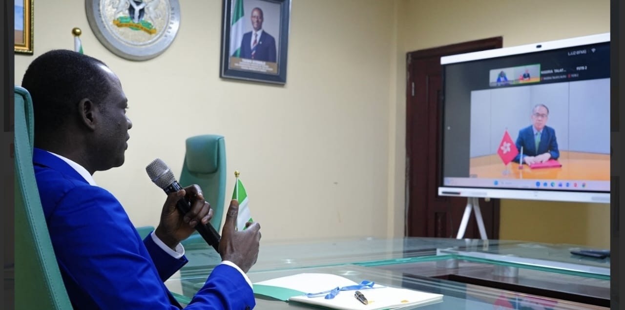 Man in a blue suit speaks into a handheld microphone during a virtual meeting, with a Nigerian flag on the desk and a video call on a large screen.