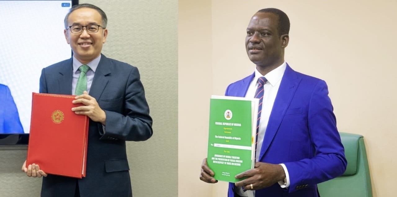 Two men in suits hold official folders: one with a red binder, the other with a green booklet, posing for a photo in a formal setting.