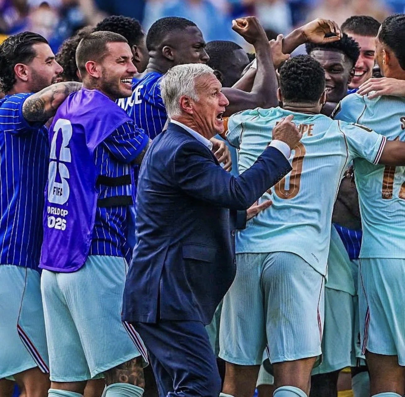 Coach in a dark suit leads a jubilant soccer team in light-blue kits during a post-match celebration on the field.
