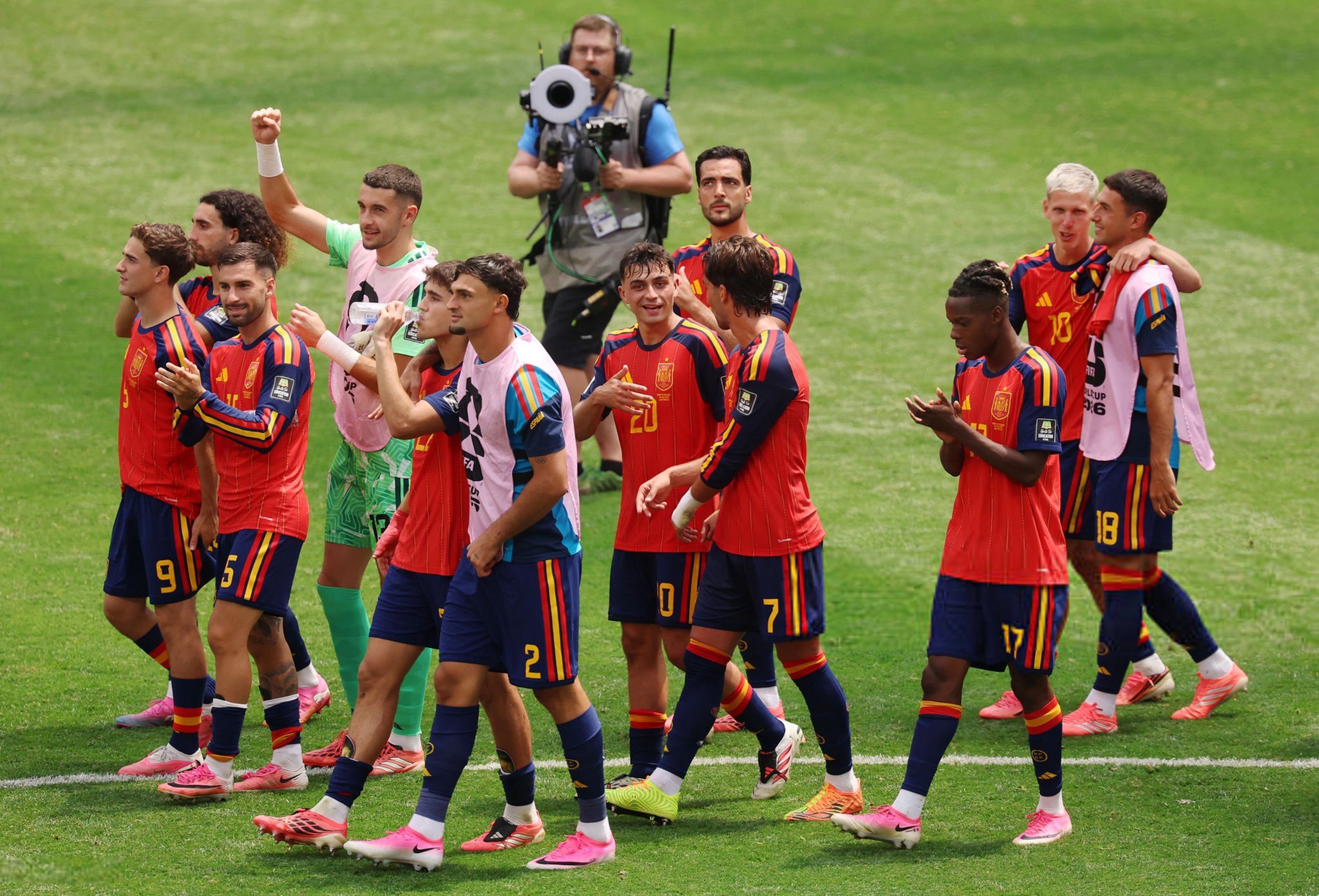 Group of soccer players in red-and-blue uniforms walking off the field, with a green-clad goalkeeper, while a cameraman films in the background on a grassy pitch.