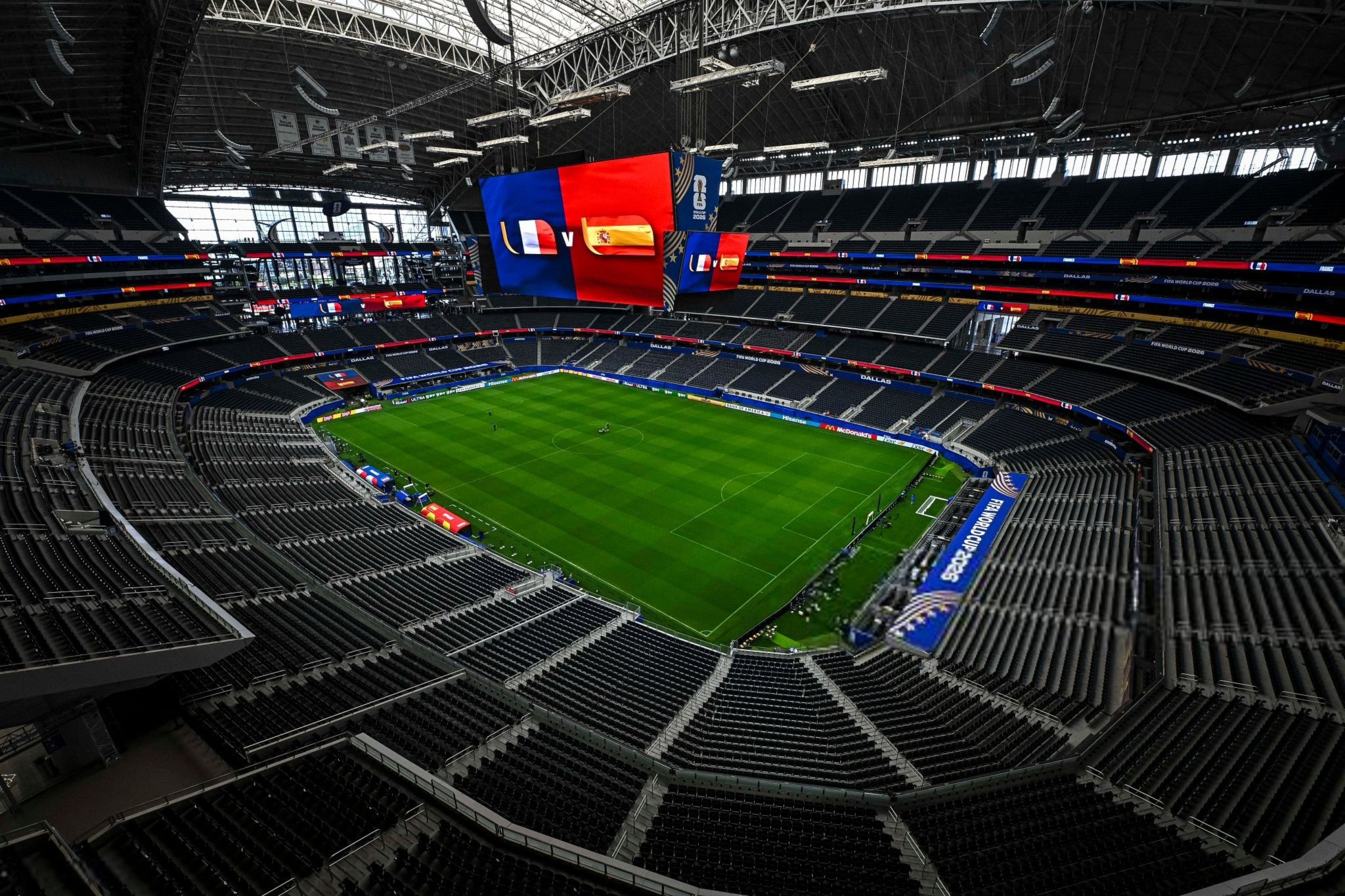 Interior of a large football stadium with a green pitch and empty black seats, a massive hanging video screen displaying blue and red panels and flags above the field.
