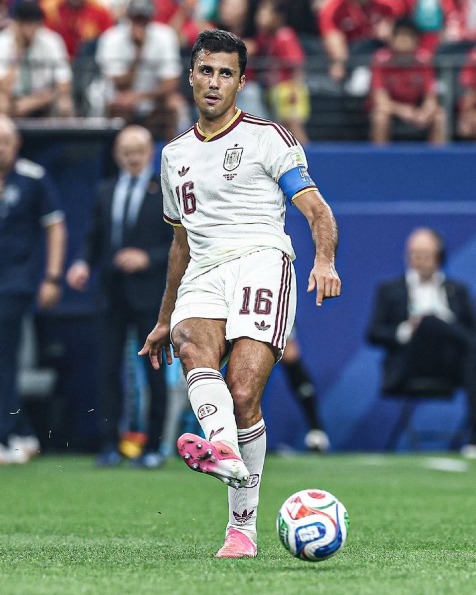 Soccer player in a white Spain jersey with number 16, kicking a ball on a green field during a match, blue captain's armband visible.
