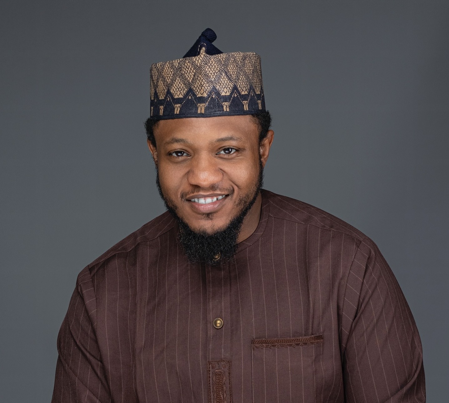 Muhammad Hadi Mutallab - THEWILL NEWS MEDIA Smiling man wearing a patterned traditional cap and brown striped shirt against a grey background