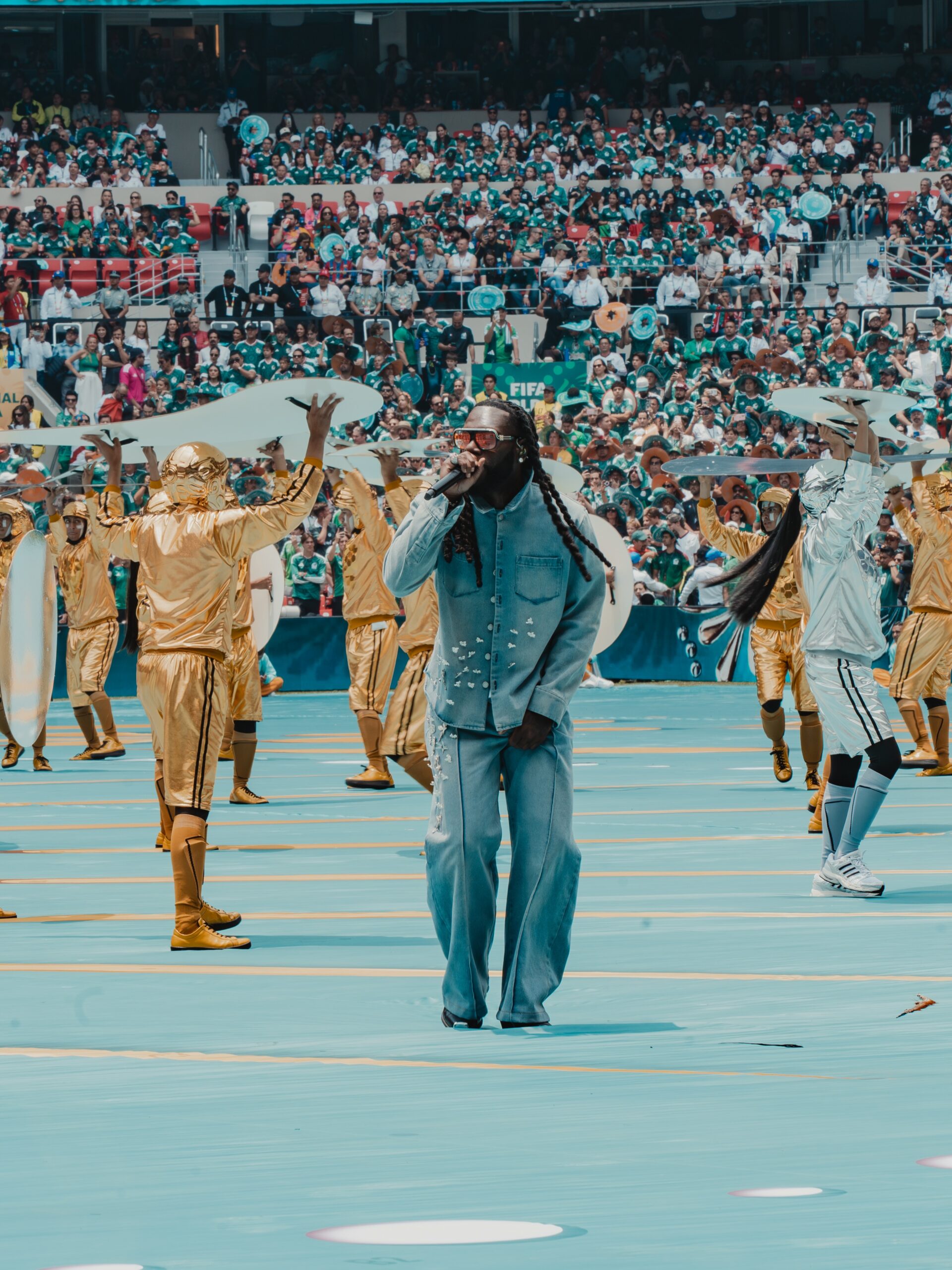 Burna Boy singing centre-stage with costumed World Cup performers behind him during the 2026 FIFA World Cup opening ceremony. Photo Credit: Burna Boy via Instagram) - THEWILL NEWS MEDIA Singer with braids performs on a turquoise stage, flanked by gold-dressed dancers as a large crowd watches from green-seated stands.