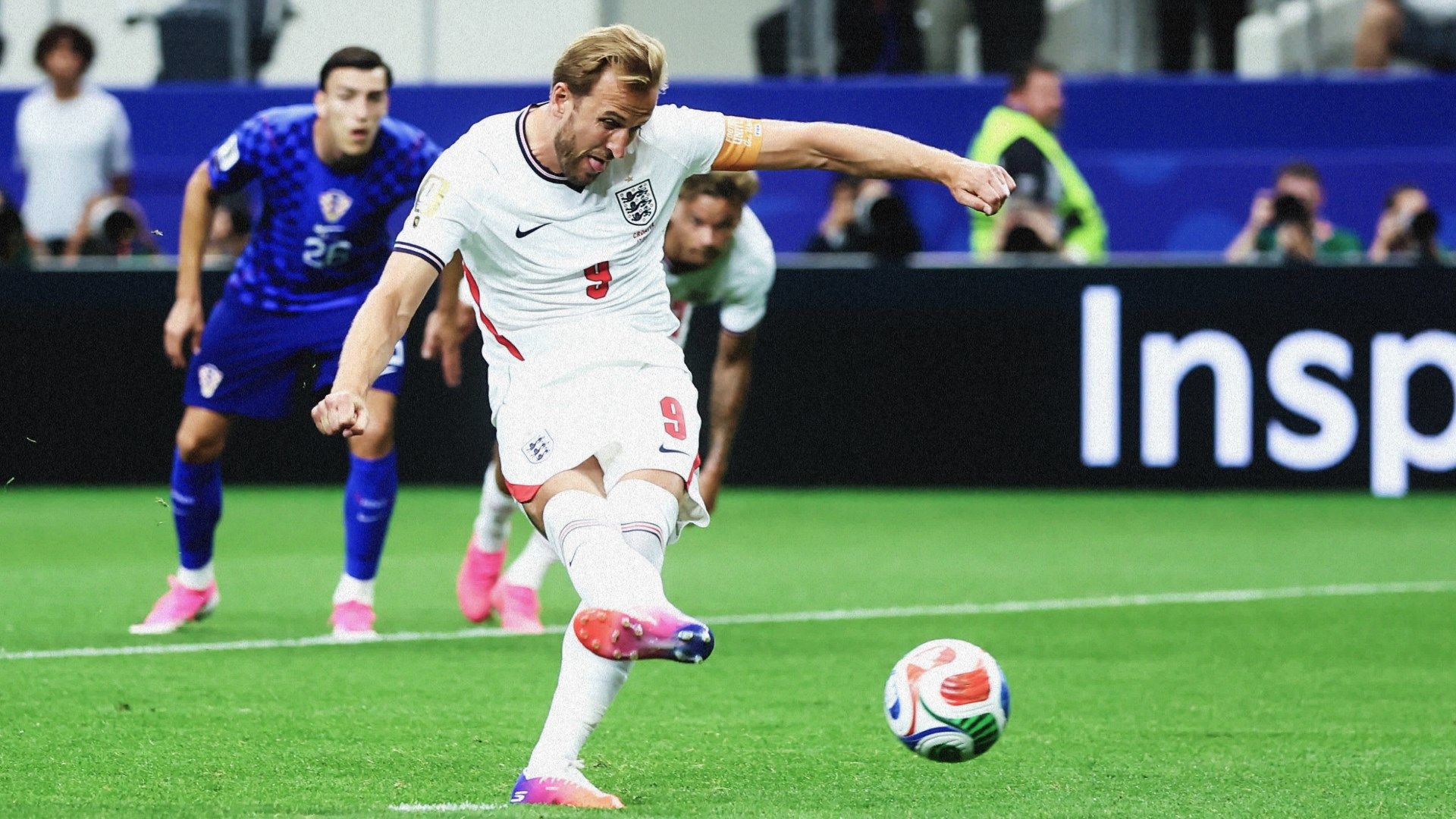 Harry Kane takes a penalty kick - THEWILL NEWS MEDIA England footballer in white kit (number 9) kicking the ball on a green pitch, with blue-clad defenders behind and photographers in the stands.
