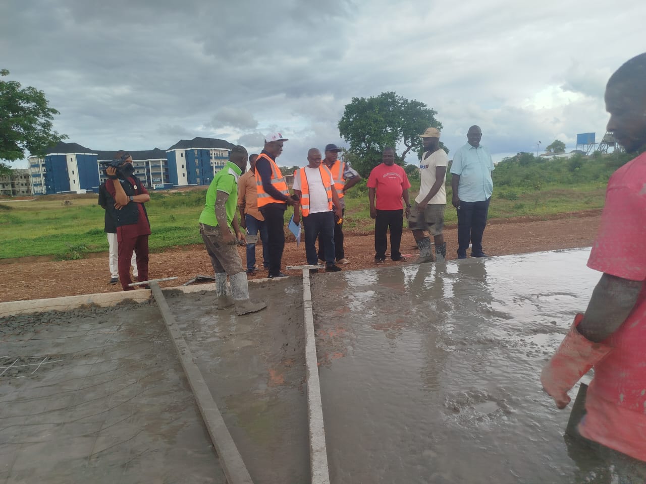 Elechi Inyima at the construction site - THEWILL NEWS MEDIA Group of workers and supervisors inspecting a freshly poured concrete slab at a construction site with muddy ground and cloudy sky.