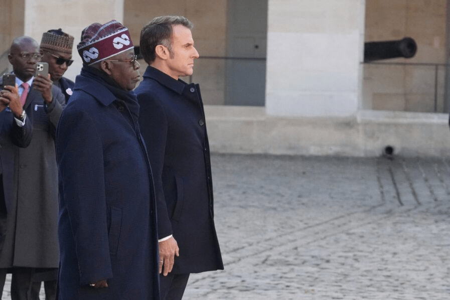France’s President Emmanuel Macron (R) and his Nigerian counterpart Bola Tinubu stand at attention during a welcoming ceremony at the Invalides in Paris, on November 28, 2024, as part of Tinubu’s state visit to France. (Photo by Michel Euler / POOL / AFP) - THEWILL NEWS MEDIA Two men in dark overcoats stand side by side outdoors, looking to the right, as photographers photograph from behind them.