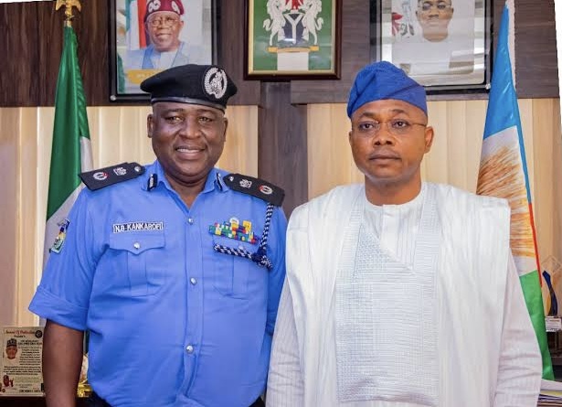 Two men pose in an office: a Nigerian police officer in blue uniform and a man in white traditional attire with a blue cap, with flags in the background.