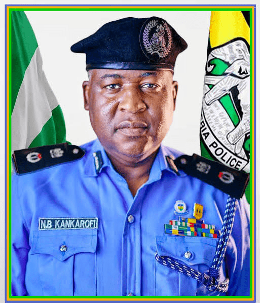Portrait of a Nigerian police officer in a blue uniform with name tag N.B. KANKAROFI, wearing a beret, medals, and flags in the background.