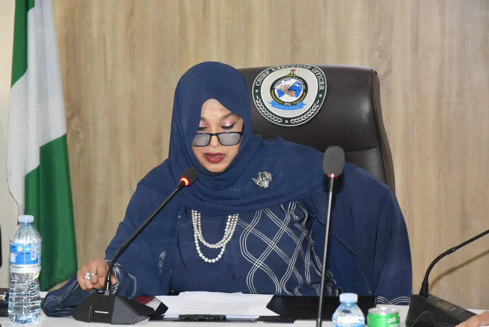 Intelligence Unit (NFIU). - THEWILL NEWS MEDIA Woman in a navy hijab and glasses speaks into a microphone at a formal meeting, with documents on the table and a seal on the chair behind her.