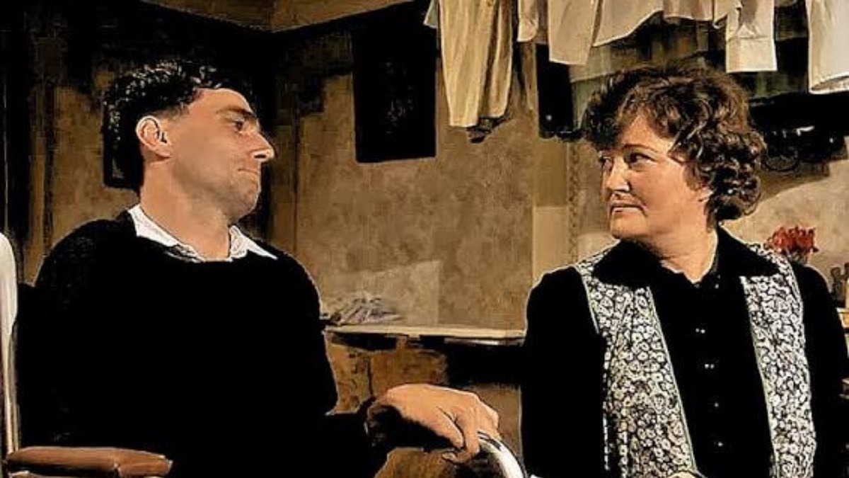 Two adults sit in a rustic room, facing each other; man on the left wears black, woman on the right has a patterned vest and dark top.