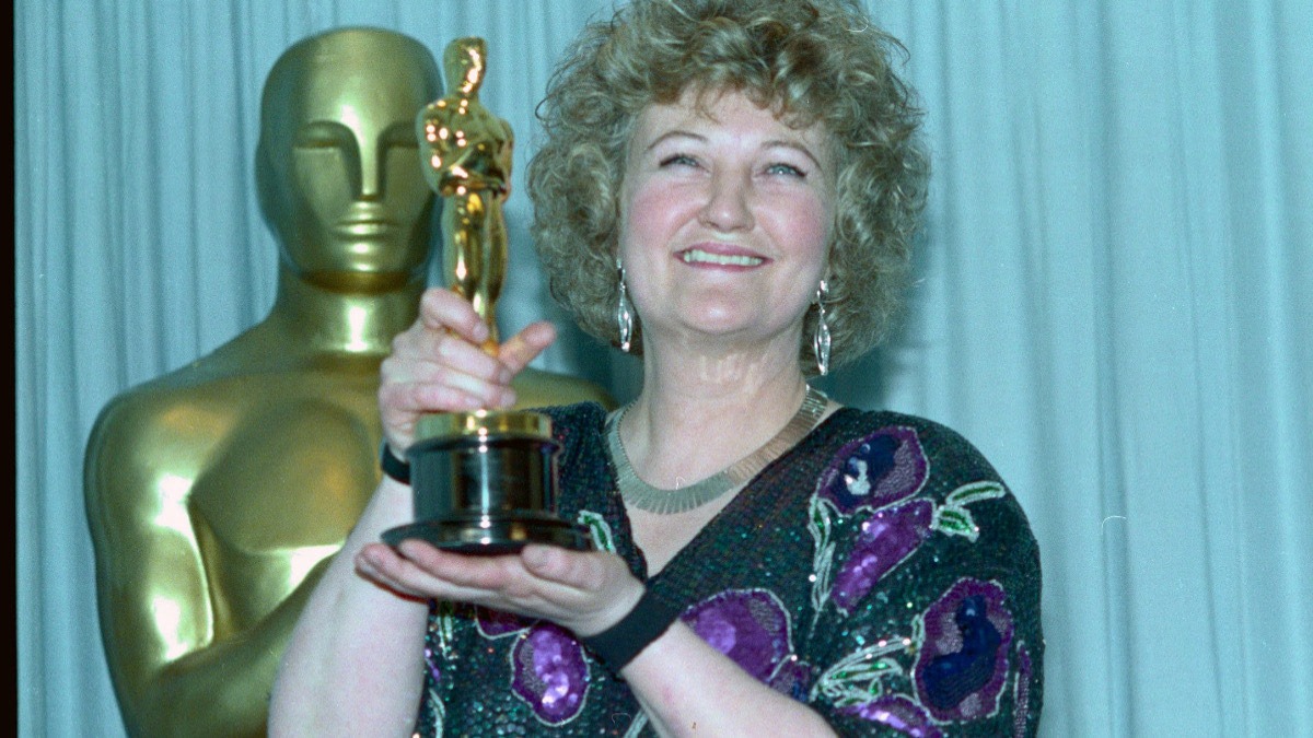 Smiling woman with curly hair holding an Oscar trophy in front of a gold statuette backdrop.