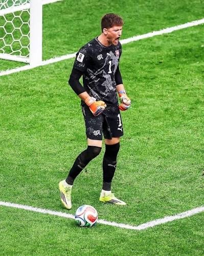 Soccer goalkeeper in black kit with orange gloves standing near a goal, ball at his feet on the green pitch.