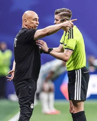 Soccer coach in black shirt argues with a neon-yellow referee on the field, pointing emphatically.
