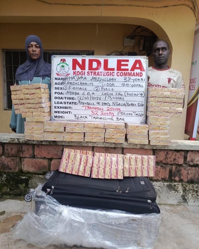 Two individuals stand beside a white NDLEA Kogi Strategic Command display board, flanked by stacked rectangular packages and a large suitcase on plastic wrap, suggesting a seized drug haul.