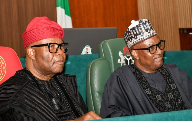 Two Nigerian men in traditional attire sit side by side at a formal conference table, with a flag and green chairs in the background.