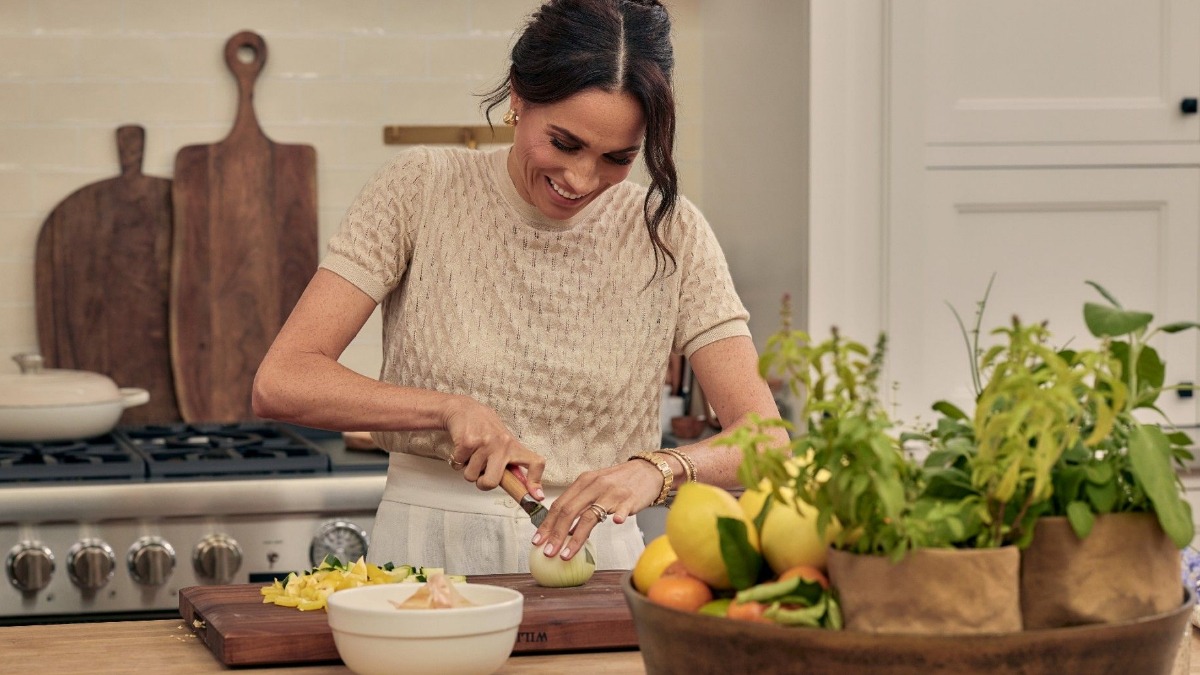 Woman chopping an onion on a wooden cutting board in a bright kitchen, with herbs and a basket of fruit nearby