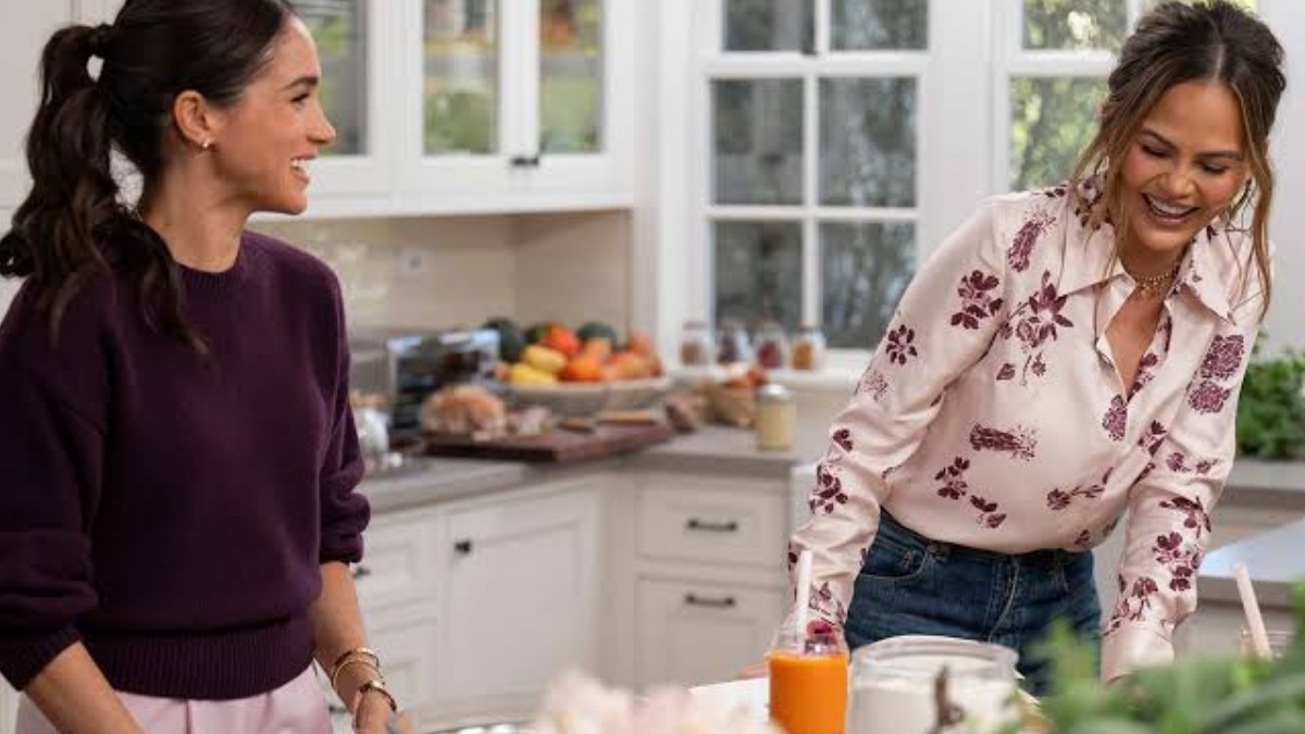 Two women smile and chat in a bright kitchen as they prep drinks, with fruit on the counter and an orange juice bottle nearby.