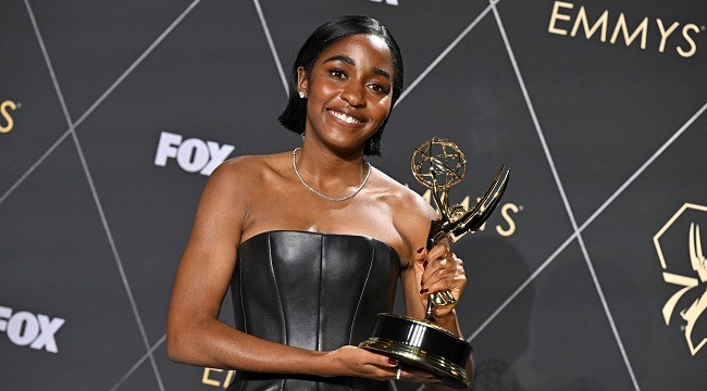 Ayo edebiri - THEWILL NEWS MEDIA Smiling woman in a strapless black dress holding an Emmy Award on the red carpet with a patterned backdrop behind her.