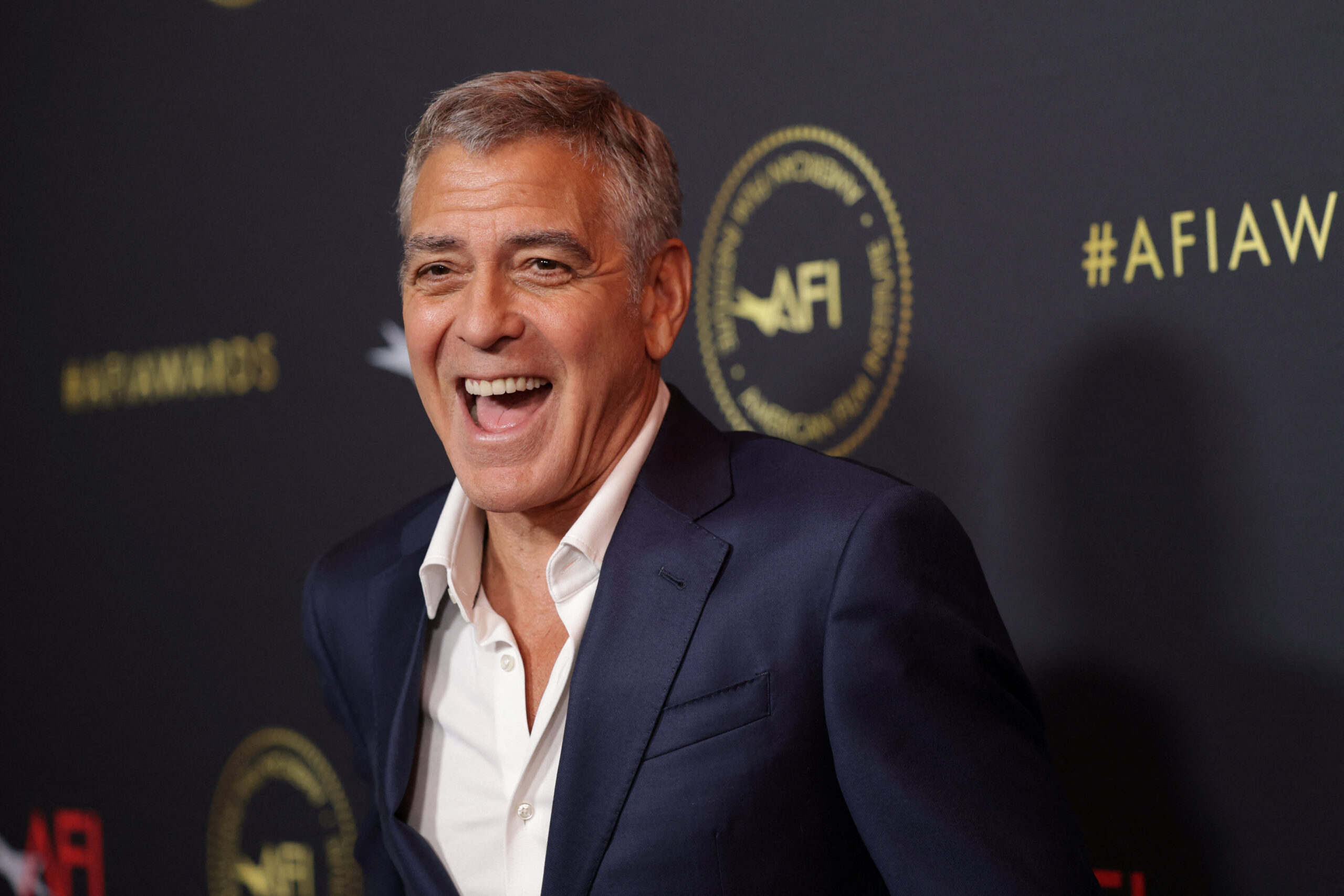 George Clooney - THEWILL NEWS MEDIA Smiling man in a navy suit photographed at an event with a dark backdrop and gold AFI branding behind him.