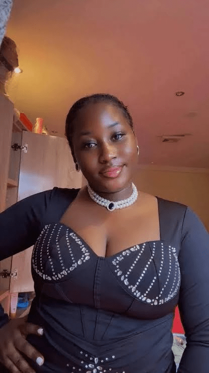 Deceased nurse- Mary Habila - THEWILL NEWS MEDIA Portrait of a woman wearing a black rhinestone-embellished dress with a pearl choker, standing indoors and smiling at the camera with one hand on her hip.
