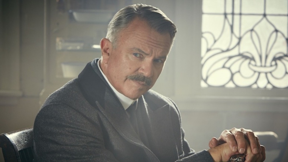 Older man in a suit sits at a desk with hands clasped, looking intently at the camera in a vintage, sunlit room
