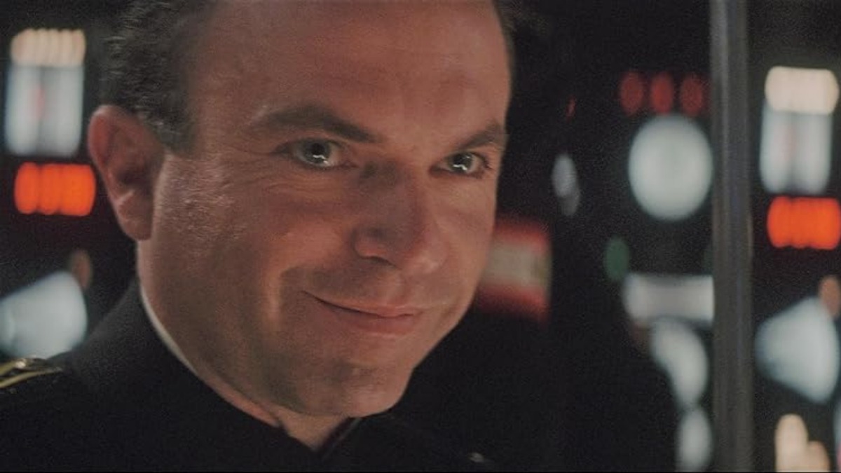 Smiling man in a dark uniform on a starship bridge with illuminated control panels behind him.